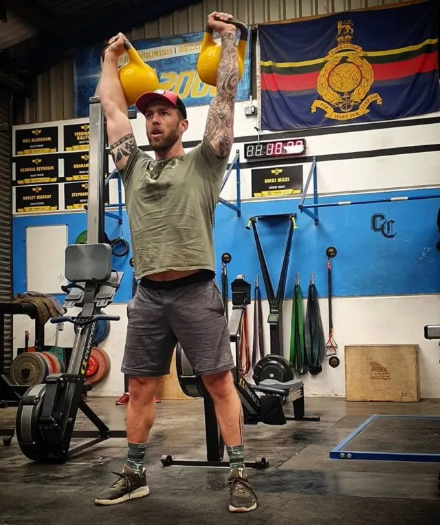 A member holding two kettlebells overhead beside the Royal Marines flag
