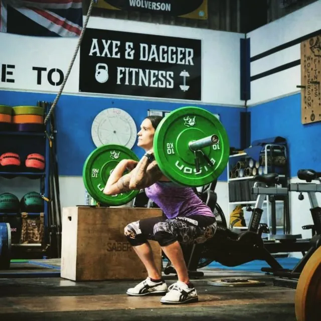 A member holding a front squat in front of the Axe & Dagger Fitness sign